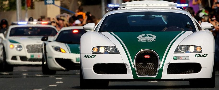A group of luxurious, expensive police cars, including a Bugatti Veyron, a Bentley, and a McLaren, drive in a parade on a city street. The cars have a green and white color scheme. The Bugatti Veyron is prominently featured in the foreground with police lights flashing.