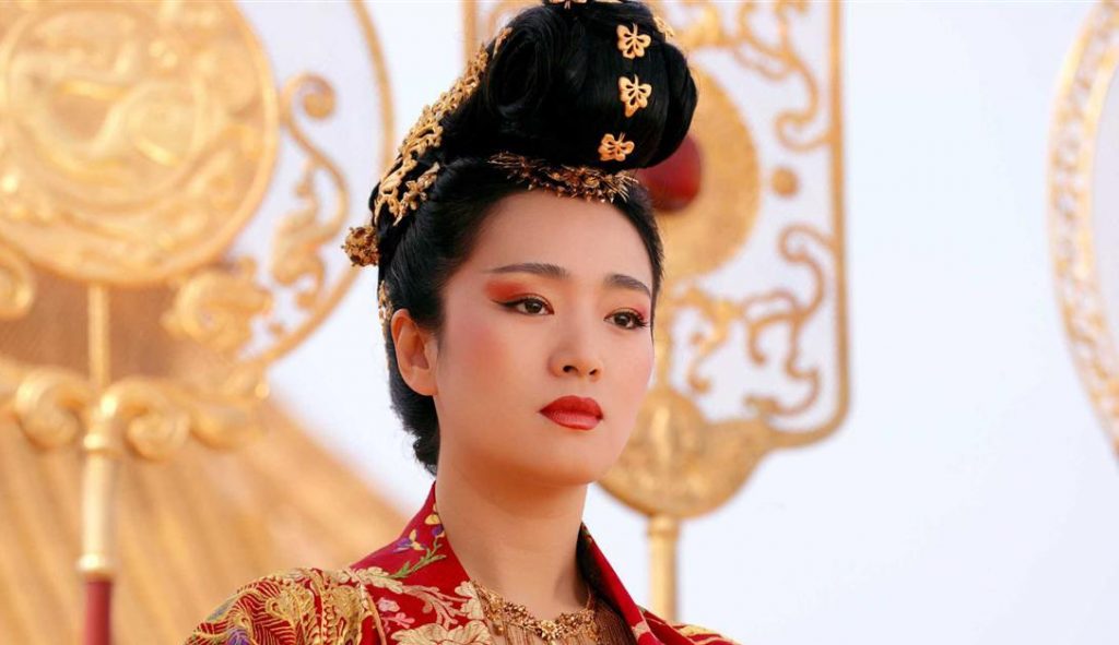 A woman dressed in traditional Asian attire wears an ornate red and gold robe and an intricate golden hairpiece adorned with flowers. Her face is decorated with dramatic red and gold makeup, set against a backdrop featuring elaborate golden decorations.